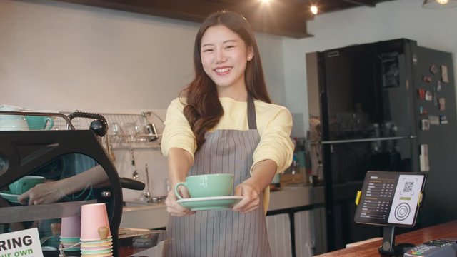 Portrait young Asian lady barista waitress holding coffee cup feeling happy at urban cafe. Asia small business owner girl in apron relax toothy smile looking to camera stand at counter in coffee shop.