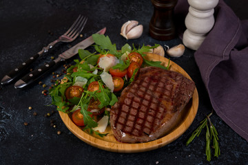 A juicy roast beef steak with fresh salad in a wooden plate stands in the center of the frame on a black concrete background. Horizontal orientation..