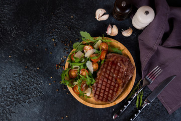 A juicy roast beef steak with fresh salad in a wooden plate stands in the center of the frame on a black concrete background. Horizontal orientation. Top view.
