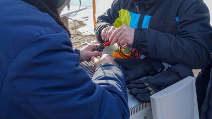 Two men in warm winter clothes use a digital multimeter to check the interface cable connector while outdoors in winter.