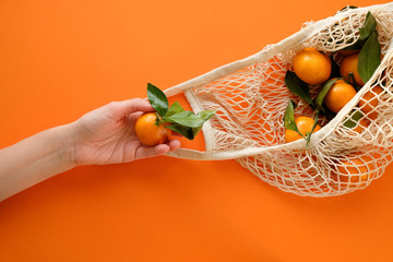 A human hand holds a string bag with tangerines on bold orange color background.