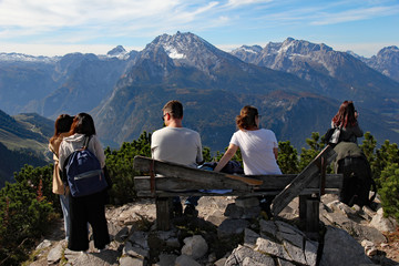 Tourists sit and stand to admire the view of the Bavarian Alps and surrounding farms and...