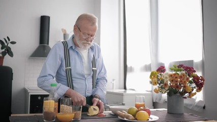 elderly attractive grandfather with a beard in glasses and takes care of his health, prepares a wholesome breakfast of fruit slices apples standing by the table