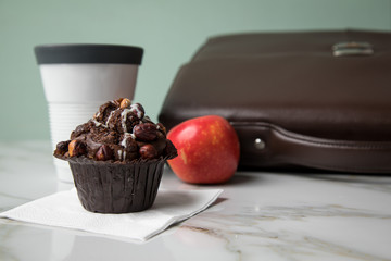 Haselnuss Muffin, Kaffee im to go Becher und Apfel als Frühstück auf dem Weg zur Arbeit mit Leder Aktentasche auf Marmor Tisch und Grün Hintergrund