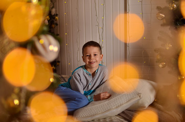 The child is sitting on the bed with a beautiful decor for Christmas. Around the baby a lot of gifts. A boy in pajamas in a beautiful interior looks at a decorated.Christmas tree. Bright photo in warm