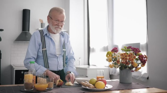 old peppy man with a beard in glasses a lover of proper nutrition prepares healthy fruity salati drinks vitamin juice, while standing at the table background of a beautiful kitchen