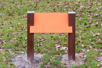 Blank orange wooden board for information and indications in the park, with a background of green grass covered by autumn leaves