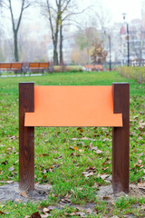Blank orange wooden board for information and indications in the park, with a background of green grass covered by autumn leaves