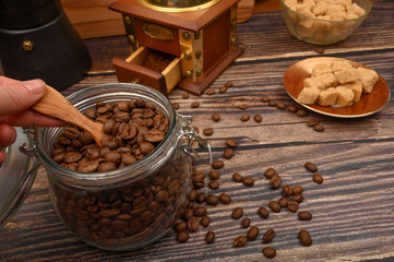 The girl's hand takes a wooden spoon of coffee beans from a glass jar, a coffee grinder, pieces of brown sugar on a wooden background. Close up.
