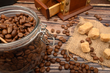 Wooden spoon in a glass jar with coffee beans, coffee grinder, pieces of brown sugar on a wooden background. Close up.