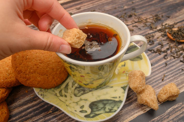 The girl's hand puts a piece of brown sugar in a mug of black tea, oatmeal cookies, tea leaves, brown sugar on a wooden background. Close up.