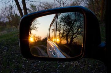 Side rearview mirror of the car with the reflection of the beautiful sunset in the forest. Close up. View of the sun through the autumn trees. Concept of family road trips and travel.