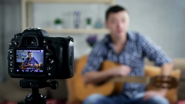 Happy man at home playing guitar. Young man working as web influencer, recording vlog and music tutorial for the Internet- Focus on camera.