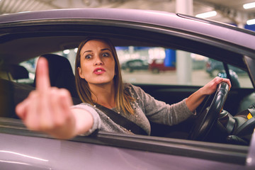 Young woman driver showing middle finger trough the window of the car mad angry furious pissed on...
