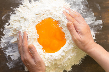 Heap flour with yolks in middle to making a dough with woman hands on wooden background
