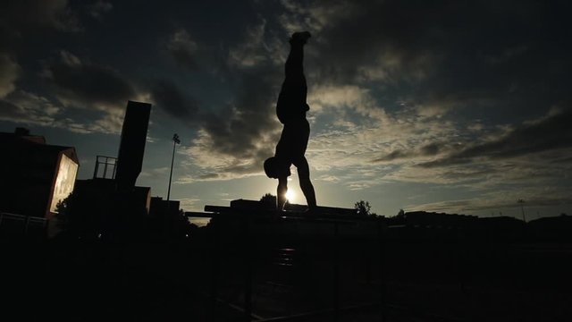Silhouette of a young man exercising push-ups on parallel bars outdoors. Bottom view. Cloudy sky with little sunshine on the background