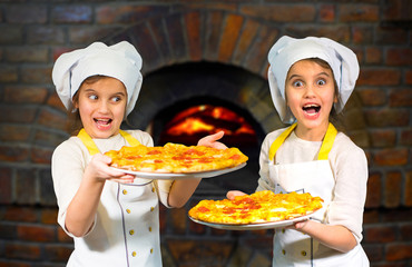 Twins children. Beautiful sisters twins girls in cook hats holding pizza on stone stove background.