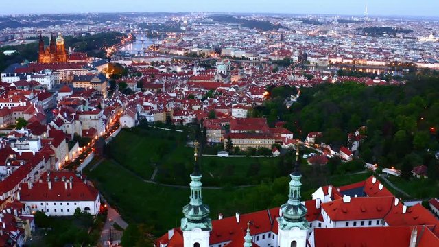 Aerial view Prague in twilight and Strahov monastery. Drone flaying backwards above roof and towers of ancient cathedral on hilltop over town. Overall picturesque panorama illuminated European city