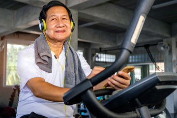 Senior Asian man in sportswear listen to music and training biking cardio at fitness gym.