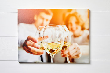 Canvas photo print on white wooden background. Stretched photo with wine glasses and happy people . Front view of colorful photography hanging on a wall