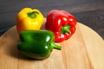 Red, green, and yellow bell peppers, cutting board on wooden background. Three sweet peppers in different colors on brown table, vegetable ingredient, healthy food