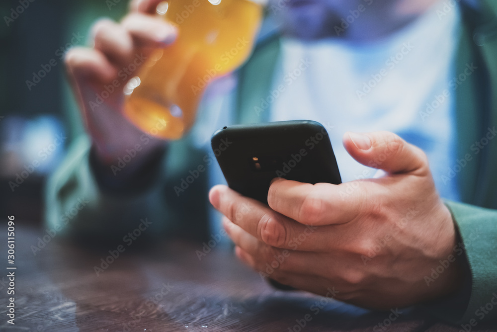 Wall mural close up of man hand hold smartphone, drinking beer and reading message at bar or pub