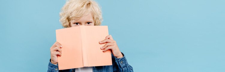 panoramic shot of cute kid obscuring face with book isolated on blue