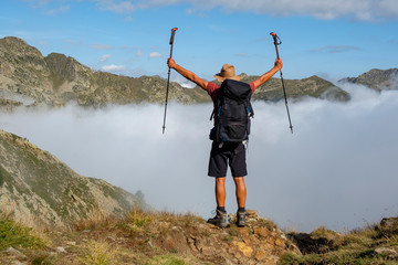 A mountain hiker with backpack, hat and shorts reaches the mountain pass in the sunshine, while...