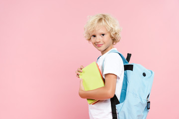 smiling schoolboy with backpack holding book isolated on pink