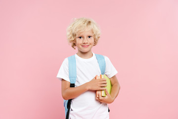 smiling schoolboy with backpack holding books isolated on pink