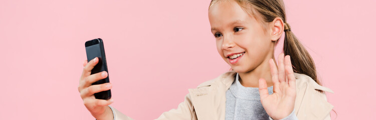 panoramic shot of smiling and cute kid waving during video chat isolated on pink