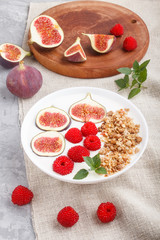 Yoghurt with raspberry, granola and figs in white plate on a gray concrete background and linen textile. side view.