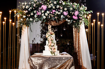 Luxurious four tiered wedding cake decorated with a lot flowers, outdoors near the festive arch, , in the evening