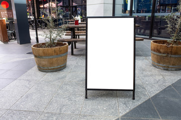 Blank white outdoor advertising stand/sandwich board mock up template. Clear street signage board placed by an outdoor dinning area of a restaurant. Background texture of standee on street.
