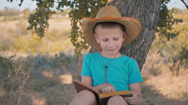A boy is reading a book sitting under a tree. A child in a straw hat reads on a hot day. Summer vacation. Rest in the shade of a tree with a book in his hands. Happy childhood.