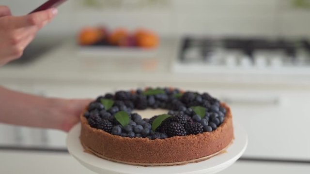 Close-up scene of a female cook who has just made a cheesecake and now has taking a photo of it on a phone.