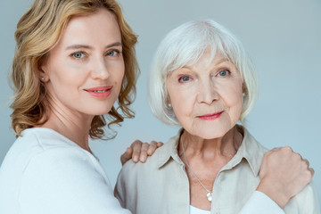smiling daughter hugging mother and looking at camera isolated on grey