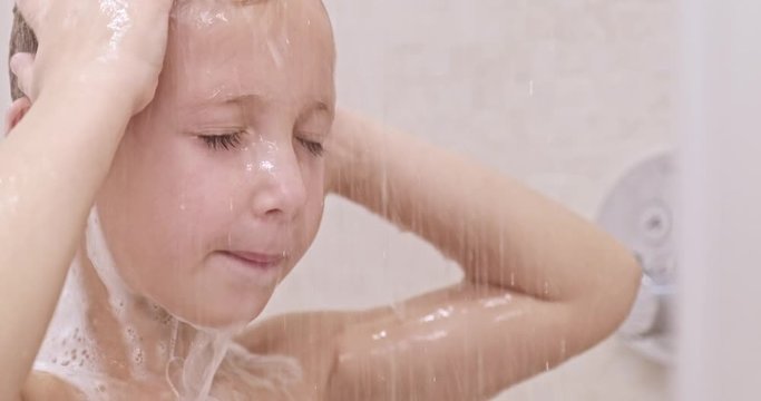 Little boy soaps hair with shampoo in the shower. Child  washes under stream of warm water. Caucasian kid takes shower in the bathroom.  European boy rinses hair with water. Slow motion.