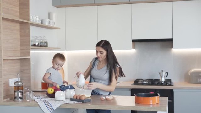 Cute little boy toddler and his mother is cooking in the modern kitchen together. Mother is mixing a dough in a bowl with a mixer and son is asking her about an apple.