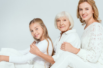 smiling granddaughter, mother and grandmother hugging isolated on grey