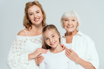smiling mother and grandmother hugging granddaughter isolated on grey