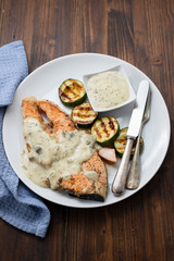 fried salmon with sauce and vegetables on white dish on brown wooden background. top view.