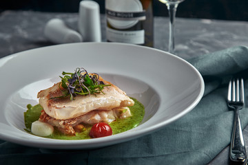 White steak of grilled fish on pillow of vegetables and yellow sauce, with tomatoes and fresh herbs. Fish on white plate with white wine on dark beautiful background. Close-up. Space