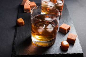 Glass of whiskey with ice and caramel candies on a black stone slate board on black background....