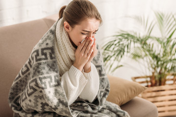 sick woman sneezing in napkin while sitting on sofa and wrapping in blanket