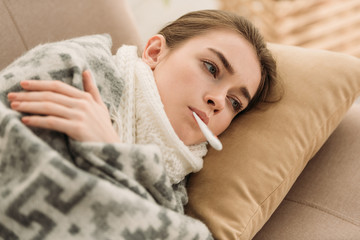 ill woman lying on sofa under blanket and measuring temperature