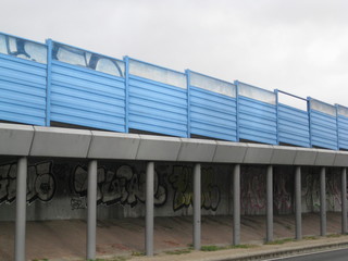 Fototapeta na wymiar vallas azules metálicas sobre escalextric hormigón, puente entre barrios con grafitis diversos en la base y columnas de pilotaje. cielo de día nublado. Paisaje urbano