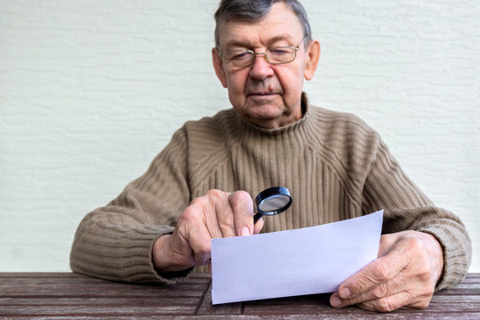 Elderly man reads fine print on white piece of paper using magnifying glass. Magnifier helps old senior