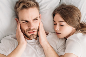 top view of exhausted man plugging ears with hands while lying in bed near snoring wife