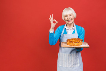 Portrait of pretty charming cheerful senior aged woman with wrinkle showing gesturing sweet homemade pie tasty bun isolated on red background, specialty of the house.